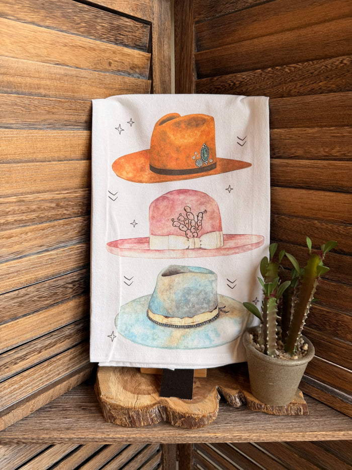 White tea towel with watercolor illustration of three hats: orange, pink, and blue