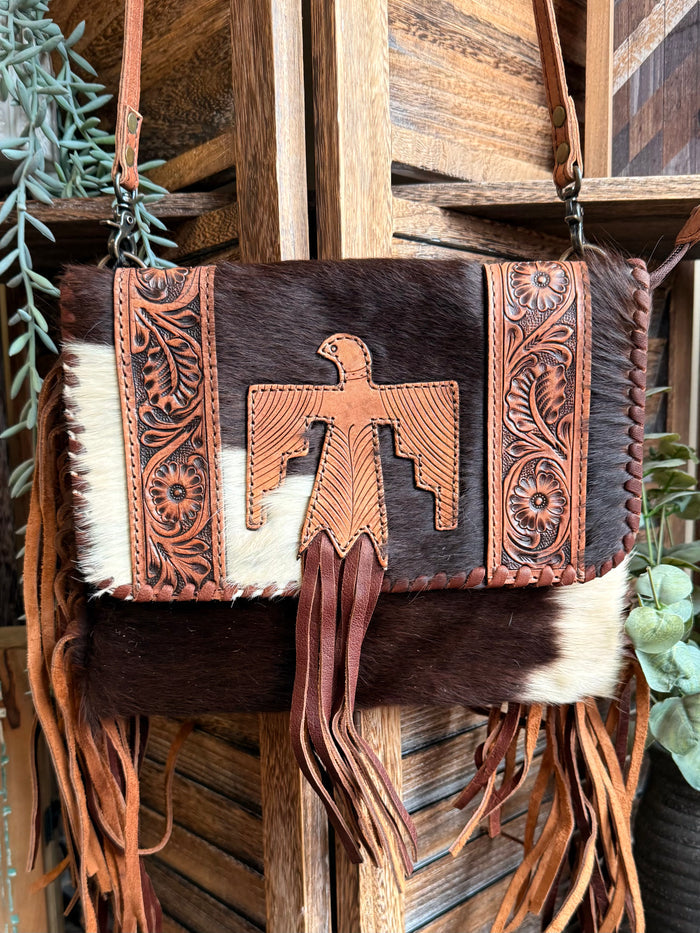 Fringed cowhide crossbody purse featuring a central tooled leather cross and brown strap