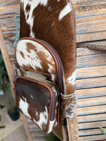 Cowhide sling bag side view with two stacked cow-print zip pockets and leather straps