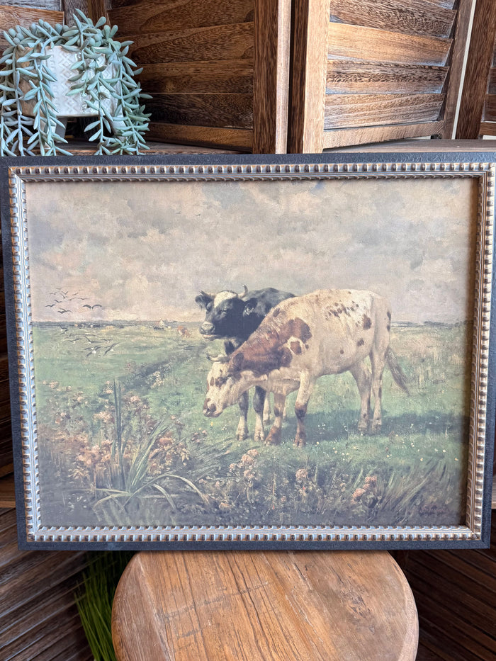 Framed prairie artwork with two cows grazing in a grassy field, blue beaded trim