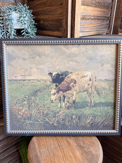 Framed prairie artwork with two cows grazing in a grassy field, blue beaded trim