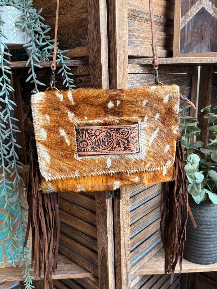 Cowhide crossbody clutch with white deer-print spots, fringe trim, and engraved clasp
