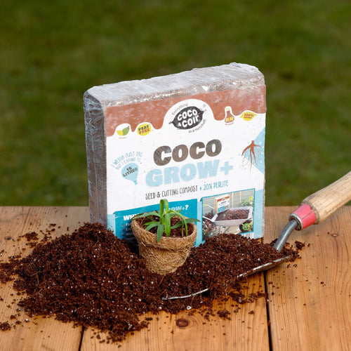 A block of coco coir potting mix next to a pot with a plant start, a shovel, and a bag of the product, all displayed on a wooden surface with soil scattered around.