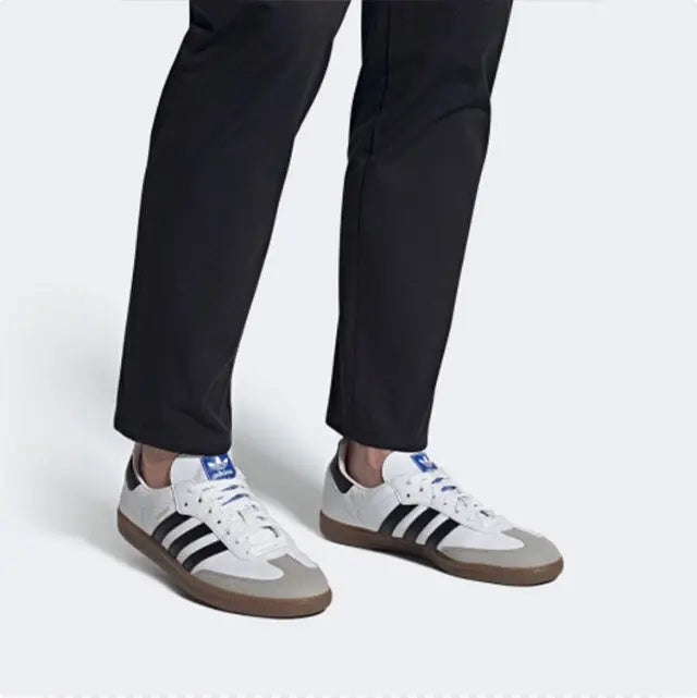 Two Adidas Samba vegan sneakers on feet with white uppers, black stripes, and gum soles.