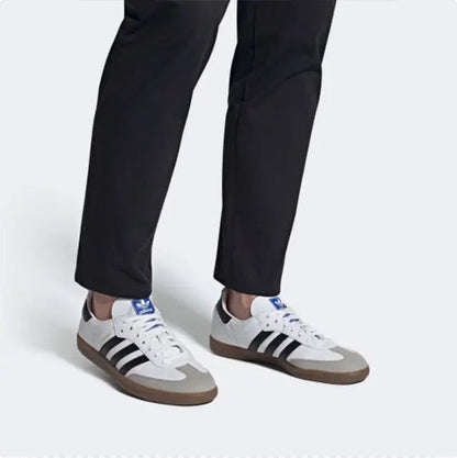 Two Adidas Samba vegan sneakers on feet with white uppers, black stripes, and gum soles.